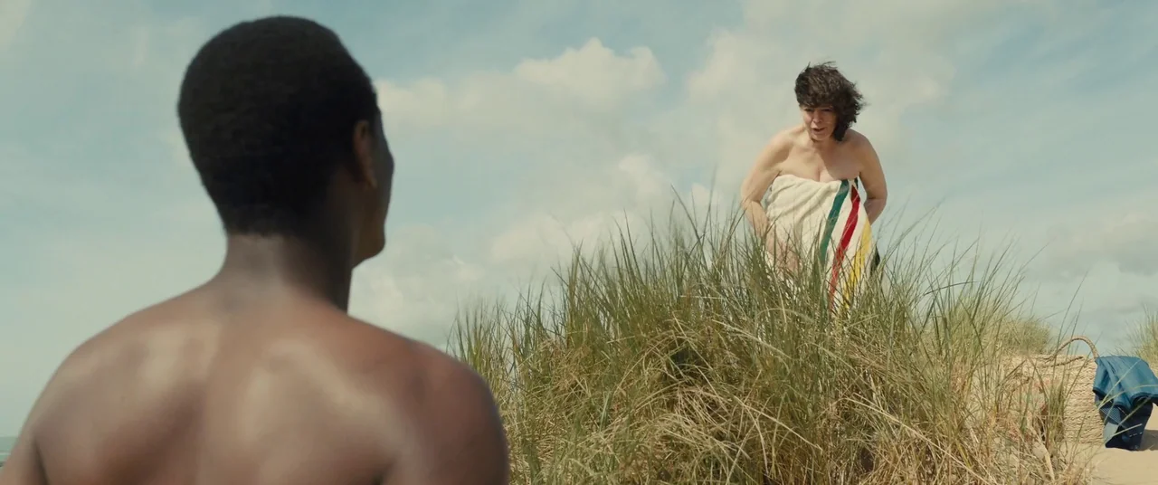 Empire of Light (2022) movie still: A quiet, intimate moment captured on a coastal dune. The composition relies on natural layers of beach grass and…