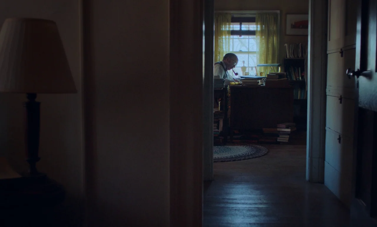 The Holdovers (2023) movie still: wide — A dimly lit room viewed through a doorway shows a cluttered home workspace. A man sits at a desk…