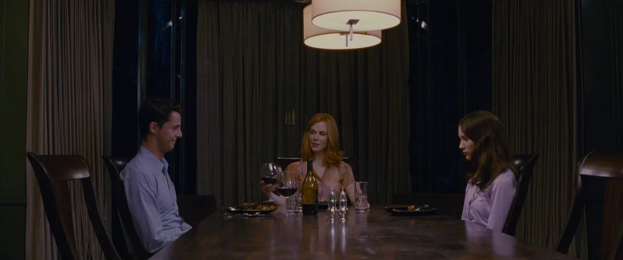 Stoker (2013) movie still: wide — A dimly lit room features a long dark wooden table beneath two overhead lamps. A man and two women sit at the…