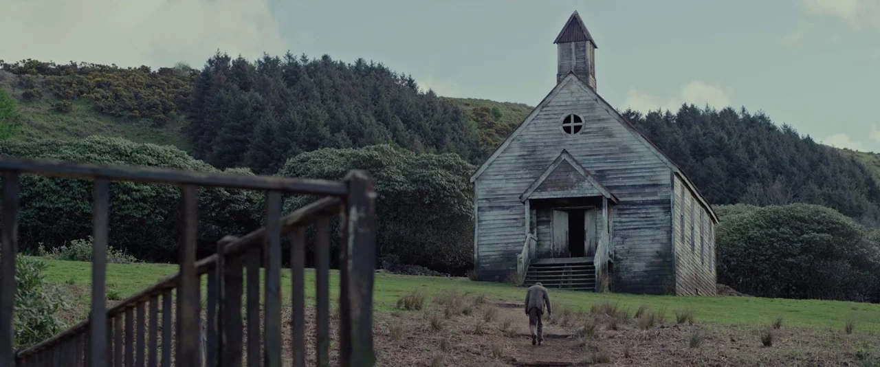 Apostle (2018) movie still: wide — An old, weathered wooden church stands on a grassy hill surrounded by thick trees. A man walks slowly along a…