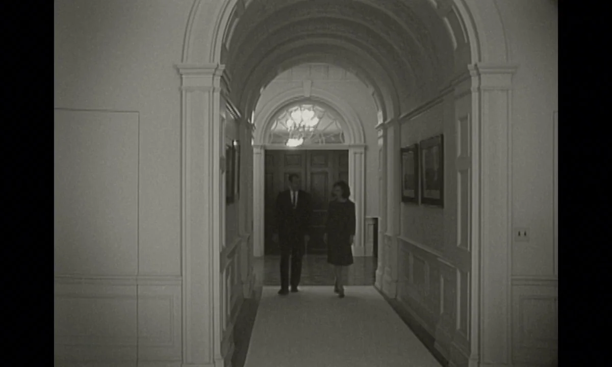 Jackie (2016) movie still: wide — A long, white-walled corridor features arched doorways and framed art on the walls. A man and a woman walk towards…