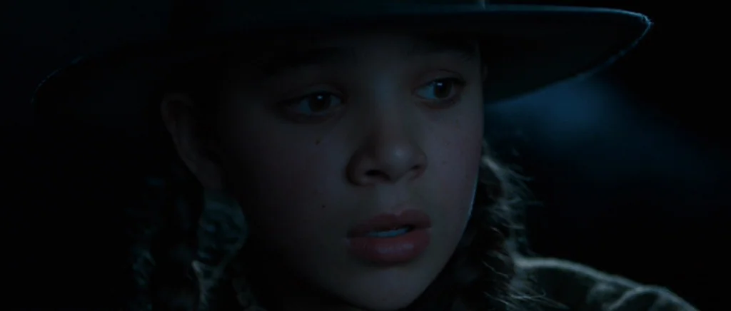 True Grit (2010) movie still: closeup — A dark night setting in a vast, featureless outdoor location. A young girl wearing a hat gazes intently at…
