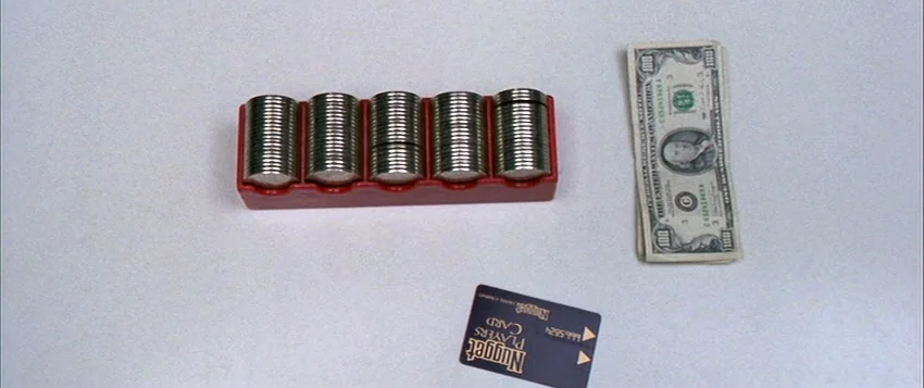 Hard Eight (1996) movie still: closeup — A top-down view shows gambling items arranged on a plain white flat surface. Stacks of metal coins in a red…
