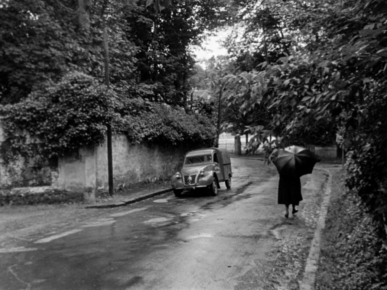 Diabolique (1955) movie still: wide — A wet, paved country road is lined with a high stone wall and thick trees. A woman with an open black umbrella…