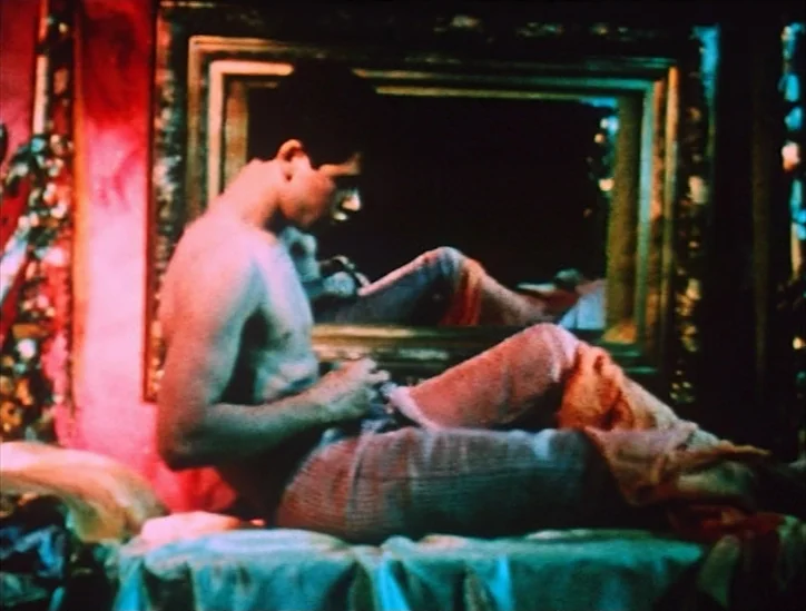 Pink Narcissus (1971) movie still: medium — A dimly lit room decorated with vivid red walls and an ornate gold-framed mirror. A shirtless man sits…