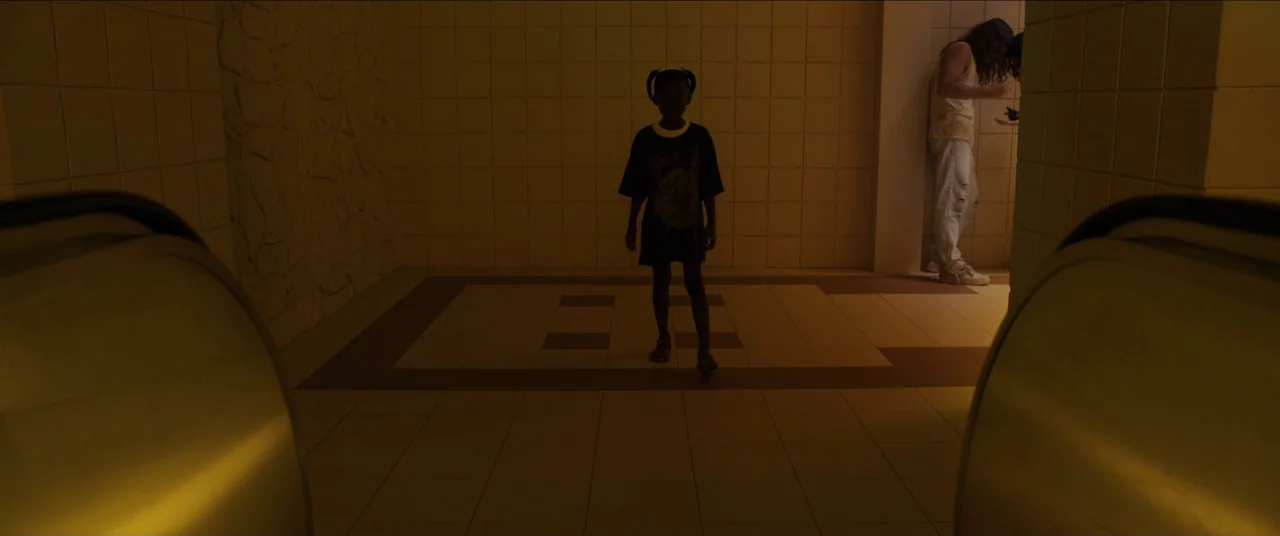 Us (2019) movie still: fullbody — A dimly lit tiled corridor features a repetitive geometric pattern on the floor surface. A young child in a dark…