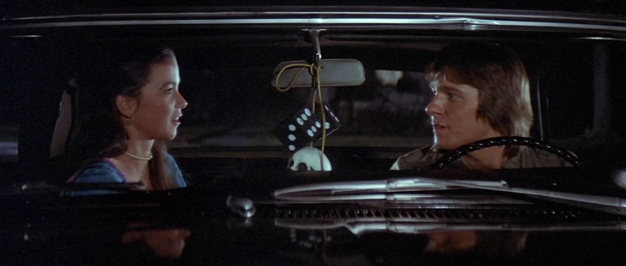 The Funhouse (1981) movie still: medium — Two individuals sit in the front seats of a moving car at night. A pair of hanging dice and a skull figure…