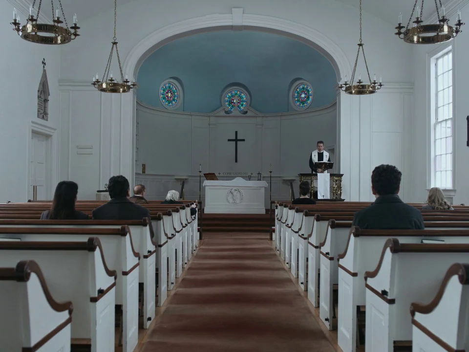 First Reformed (2017) movie still: wide — A white interior of a church with rows of wooden pews facing a pulpit. A minister stands behind the…
