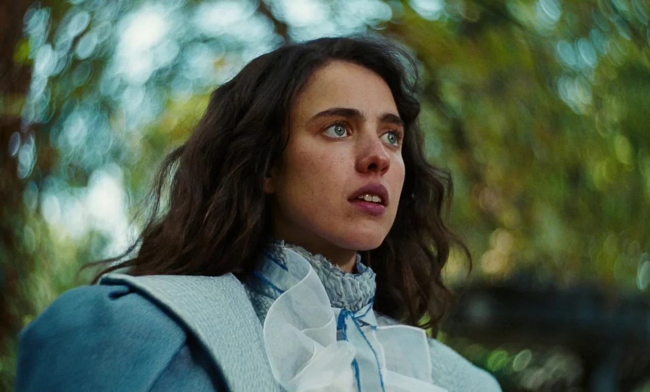 Poor Things (2023) movie still: closeup — A woman stands in a lush wooded area with blurred foliage in the background. She turns her head and looks…