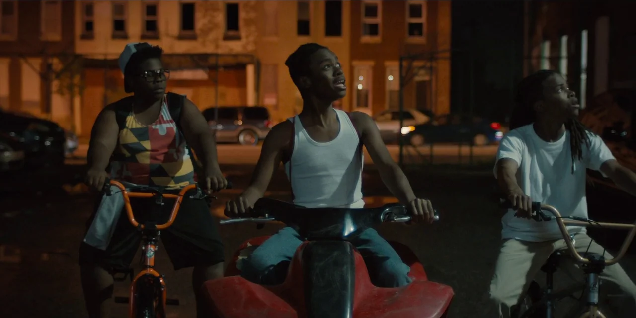 Charm City Kings (2020) movie still: medium — An urban parking lot at night with red brick buildings in the background. Three young men are…