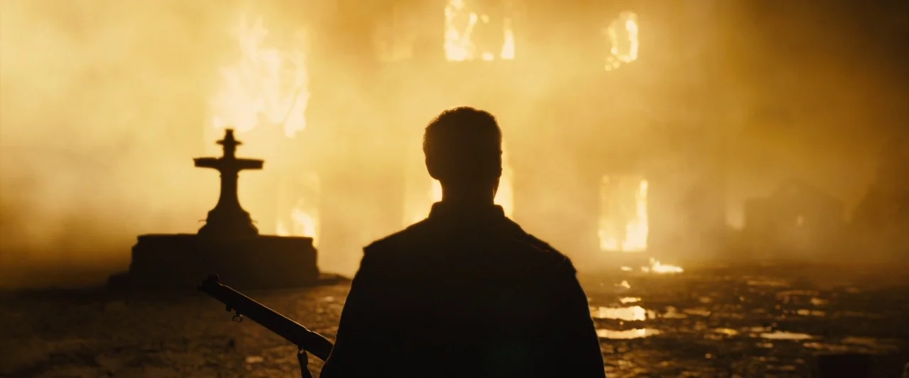 1917 movie still: medium — A destroyed village square at night is filled with dense smoke and glowing fire. A lone soldier stands in the foreground…