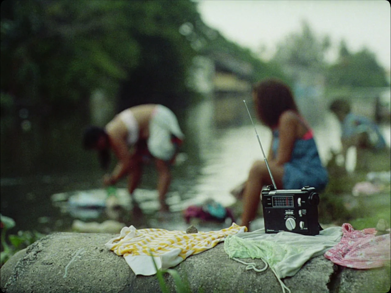 Guava Island (2019) movie still: medium — A grassy riverbank features several pieces of clothing spread out on a concrete barrier. Two women occupy…