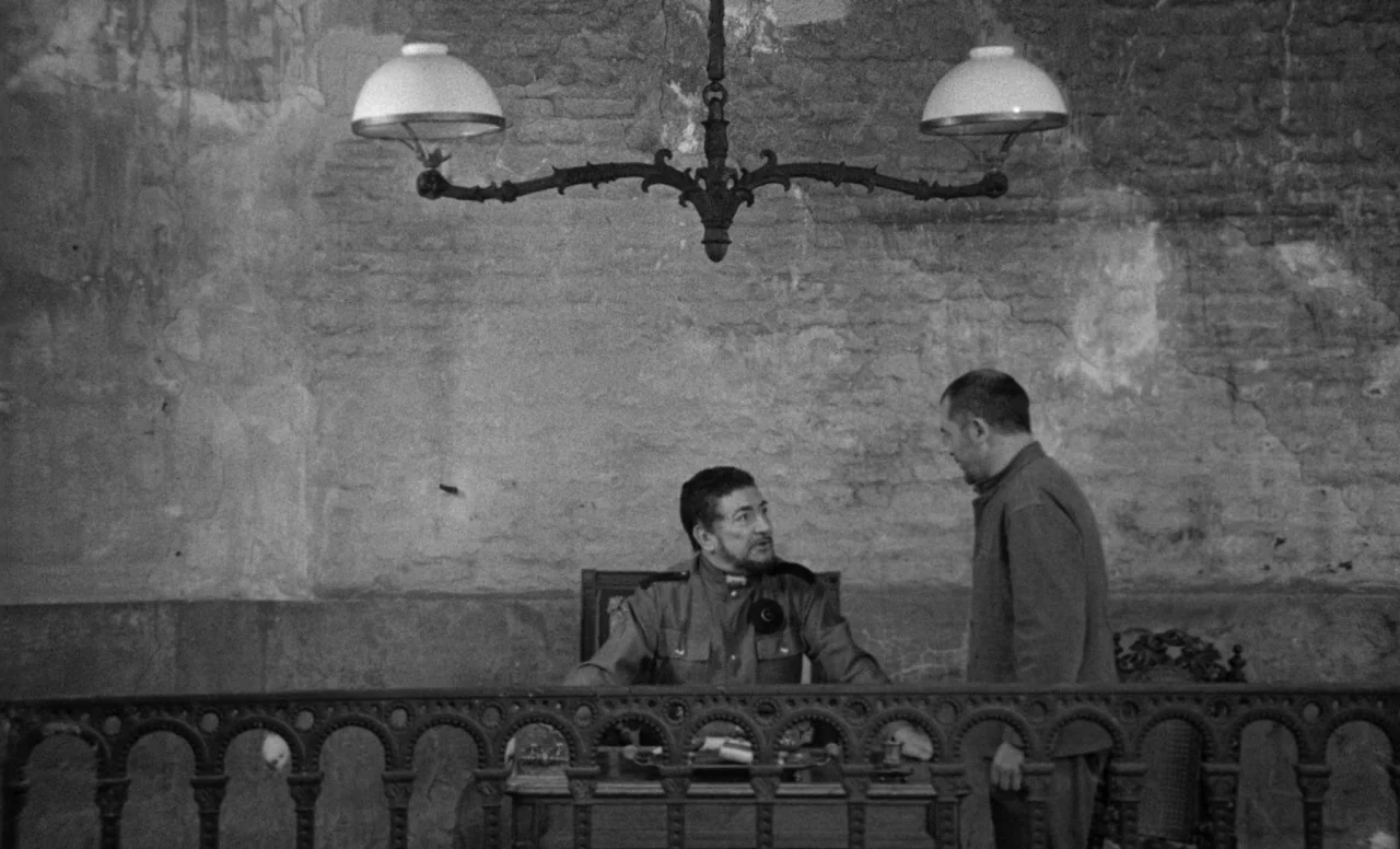 Goto, Island of Love (1969) movie still: medium — A stone-walled interior space features a desk separated by an ornate wrought-iron railing. An…