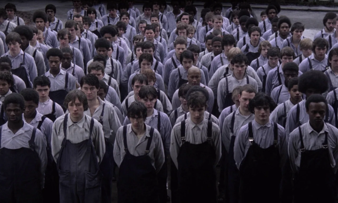 Scum (1979) movie still: wide — A large crowd of men stands in uniform rows outdoors on a concrete ground. Each individual wears an identical shirt…