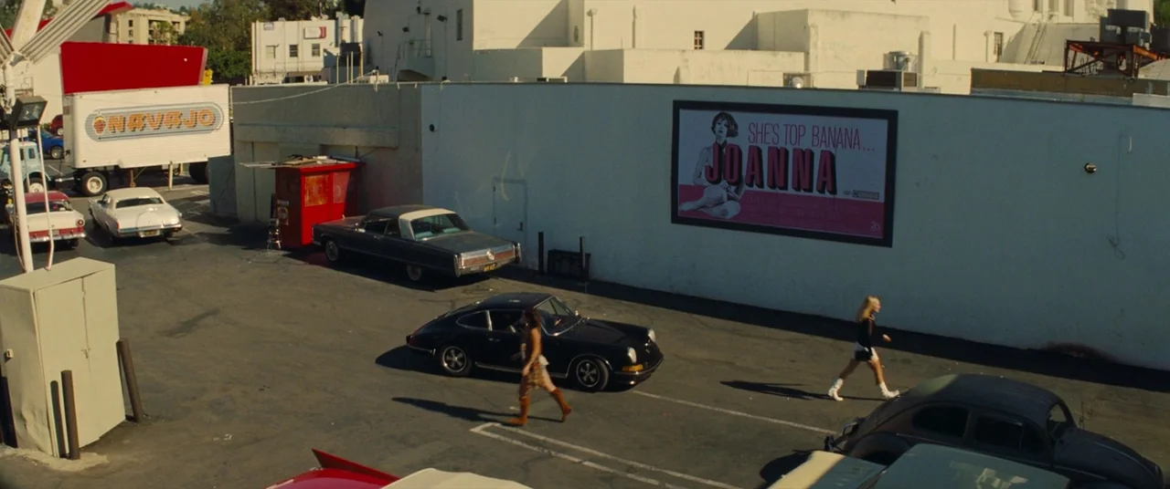 Once Upon a Time… In Hollywood (2019) movie still: wide — A sunlit parking lot features a large white wall displaying a movie poster. Two women walk…
