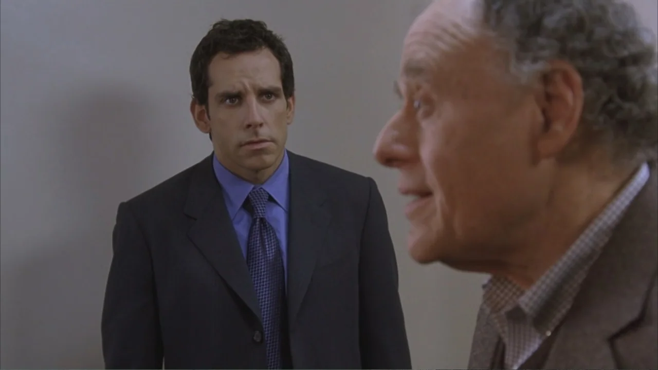 Along Came Polly (2004) movie still: medium — An interior office space with a plain, light-colored wall background. A younger man in a suit looks…