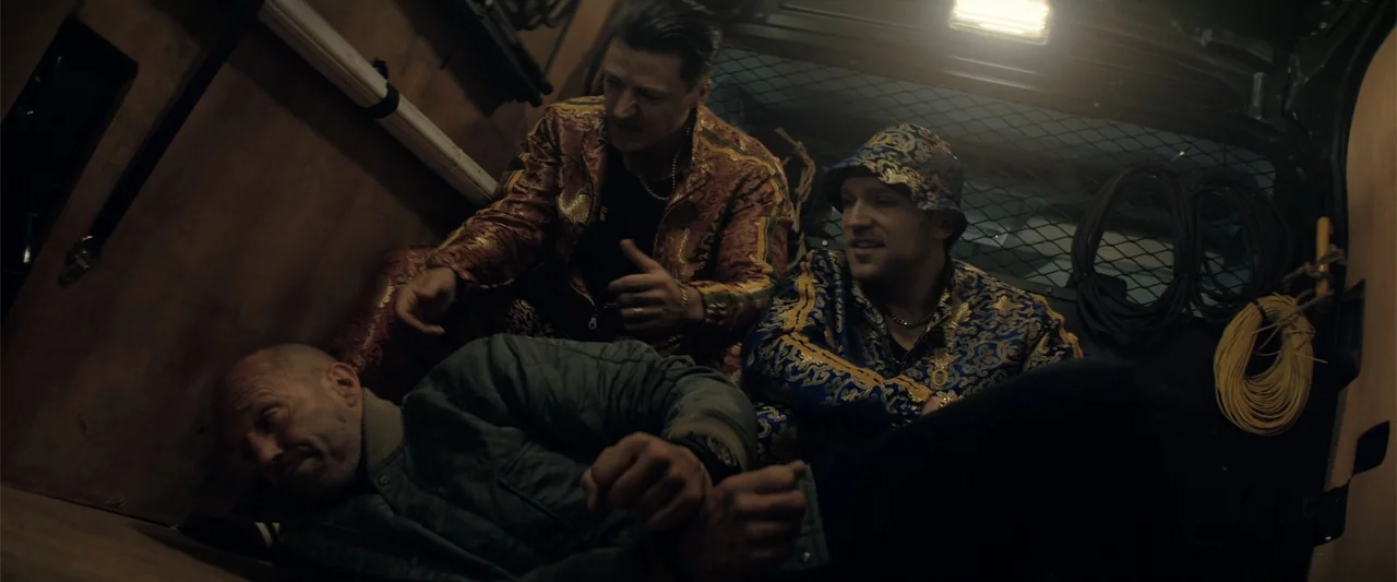 A Working Man (2025) movie still: medium — The cramped, dimly lit metal cargo space of a van contains three men. Two men sit together while looking…