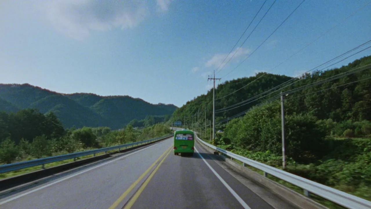 Riceboy Sleeps (2022) movie still: wide — A rural highway winds through dense green hills under a bright daytime sky. A small green bus drives away…