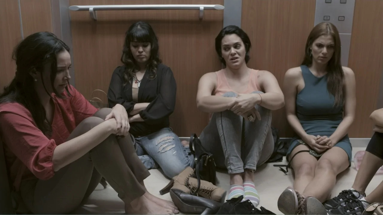 Locas y atrapadas (2014) movie still: medium — A modern elevator interior with wood-paneled walls and a metal handrail at top. Four women sit on the…