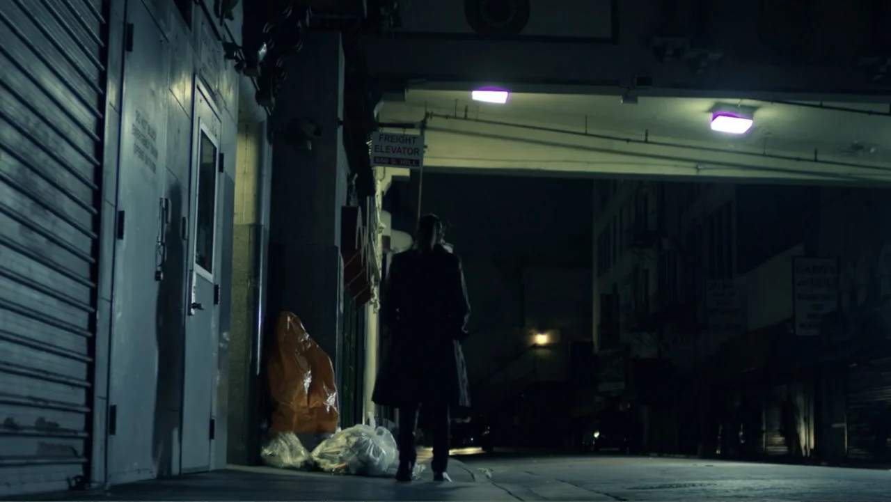 Entre las sombras (2025) movie still: fullbody — An urban alleyway features a metallic door on the left and a freight elevator sign. A solitary…
