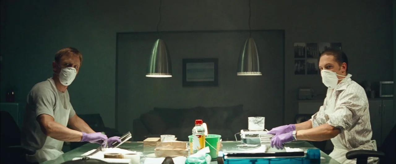 Layer Cake (2004) movie still: wide — A dark, dimly lit industrial room contains a central glass table with equipment. Two men in protective gear…