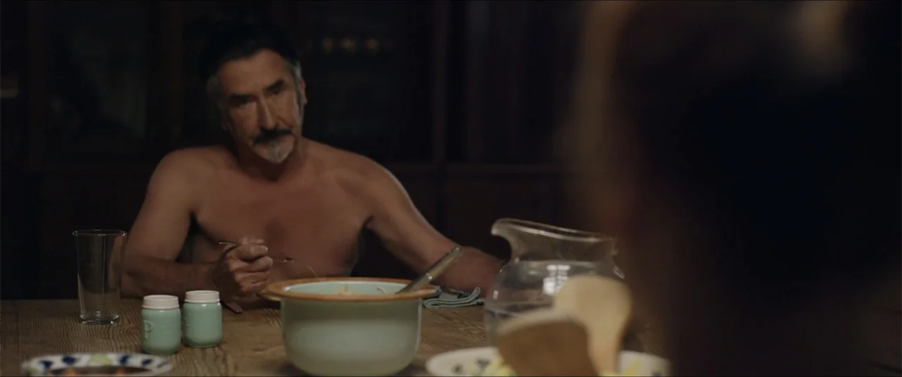 Captive (2020) movie still: medium — A dimly lit dining room features a rustic wooden table with simple food containers. A shirtless middle-aged man…