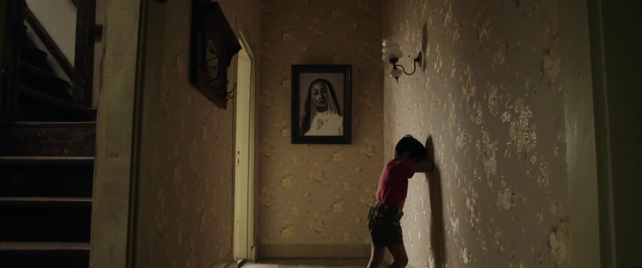 Satan’s Slaves (2017) movie still: medium — A narrow domestic corridor features floral vintage wallpaper and a framed portrait on display. A young…