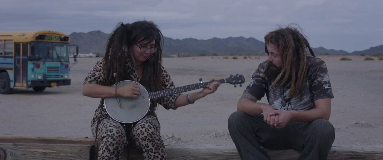 Nomadland (2020) movie still: medium — Two individuals sit on a weathered wooden beam in a vast, arid desert landscape. The woman plays a banjo for…