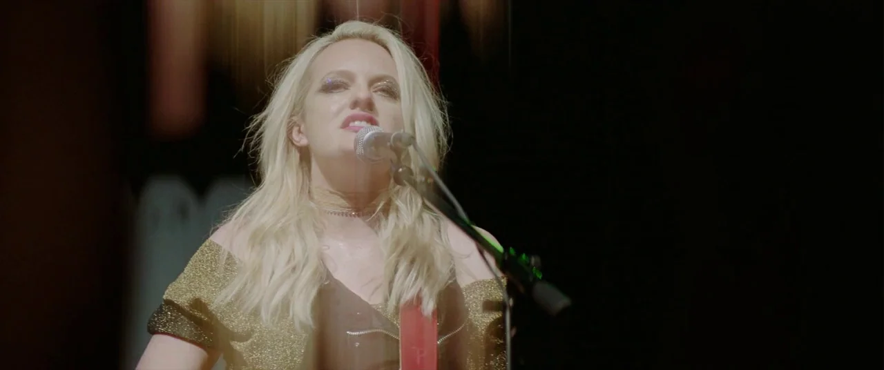 Her Smell (2018) movie still: medium — A dimly lit indoor concert venue provides a stark backdrop for a live performer. A blonde woman in a gold top…