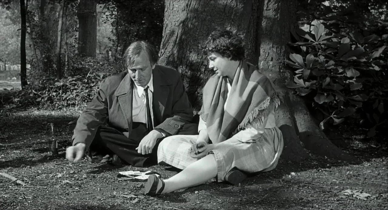 The Bohemian Life (1945) movie still: medium — A grassy area beneath a large tree with thick roots in a public park. A man and a woman sit facing…