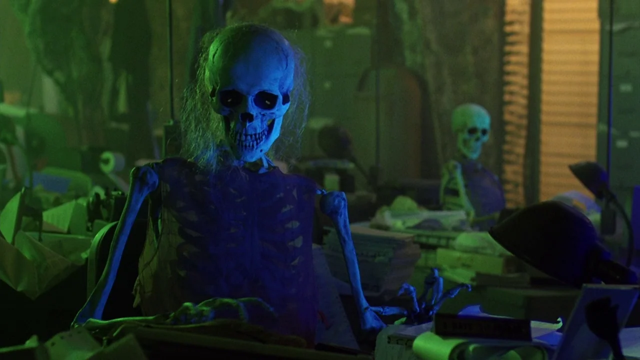 Beetlejuice (1988) movie still: medium — A dark, cluttered workspace is filled with piles of papers and vintage desk equipment. Two skeletons are…