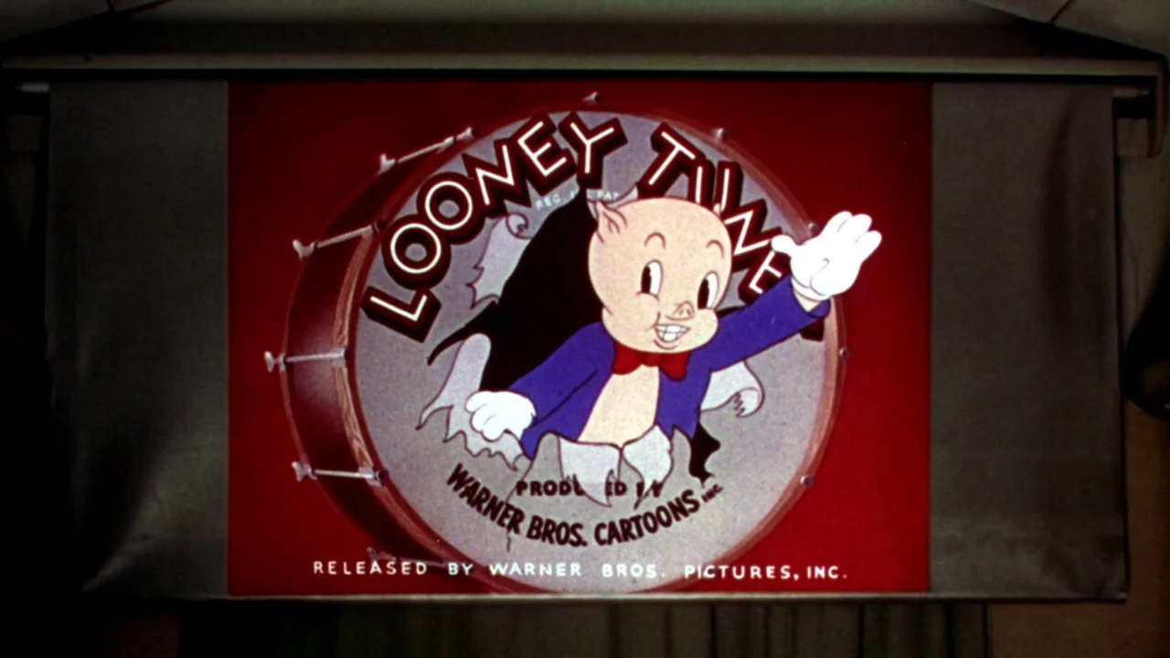 What’s Up, Doc? (1972) movie still: closeup — A cartoon title card is displayed on a wide white projection screen. The character Porky Pig pops…