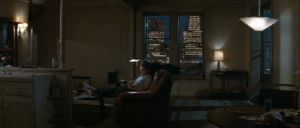 Basic Instinct (1992) movie still: wide — A dimly lit room with an open-plan layout and a large window looking onto a city at night. A man sits in a…