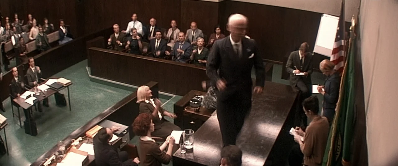 Dancer in the Dark (2000) movie still: wide — A high-angle view shows a courtroom with wooden benches and a green floor. An elderly man in a dark…