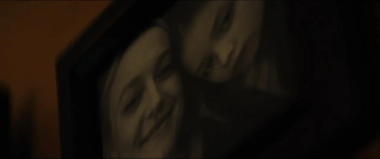 Vicious (2025) movie still: closeup — A dimly lit room holds a dark framed portrait of two young girls. The photograph is positioned at a sharp…