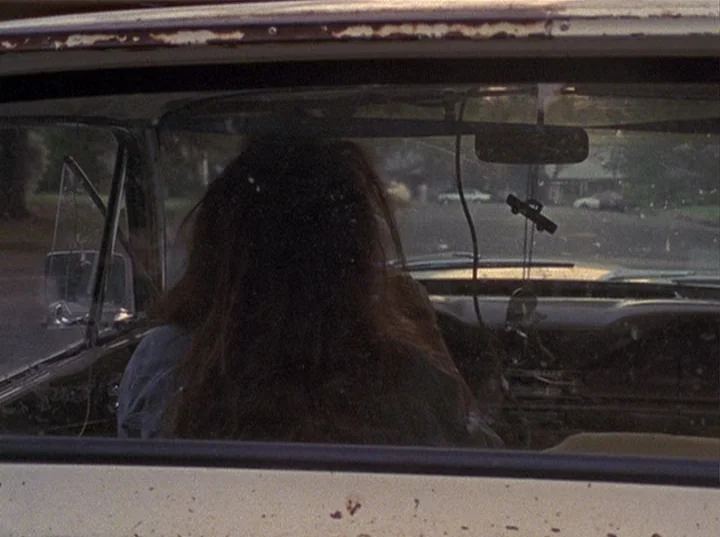 Slacker (1991) movie still: A quiet, contemplative shot of a person sitting in the driver's seat of an older car, framed from the backseat. The soft…