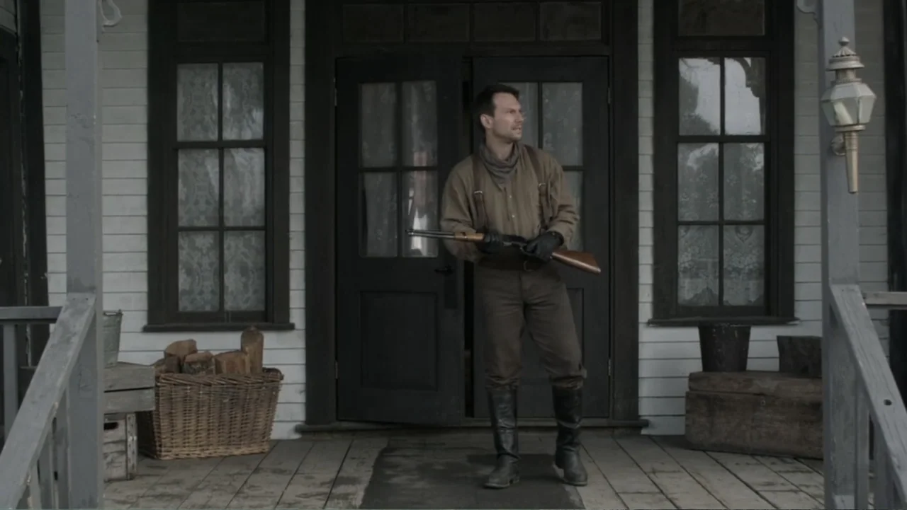 Dawn Rider (2012) movie still: fullbody — A wooden porch of a 19th-century frontier building with a basket of firewood. A man wearing period…