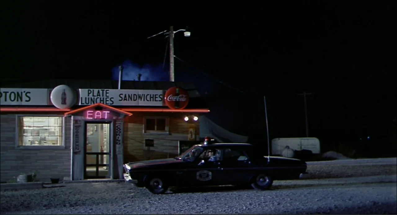 In the Heat of the Night (1967) movie still: wide — A single-story building with wood-paneled siding and glowing signage stands at night. A parked…