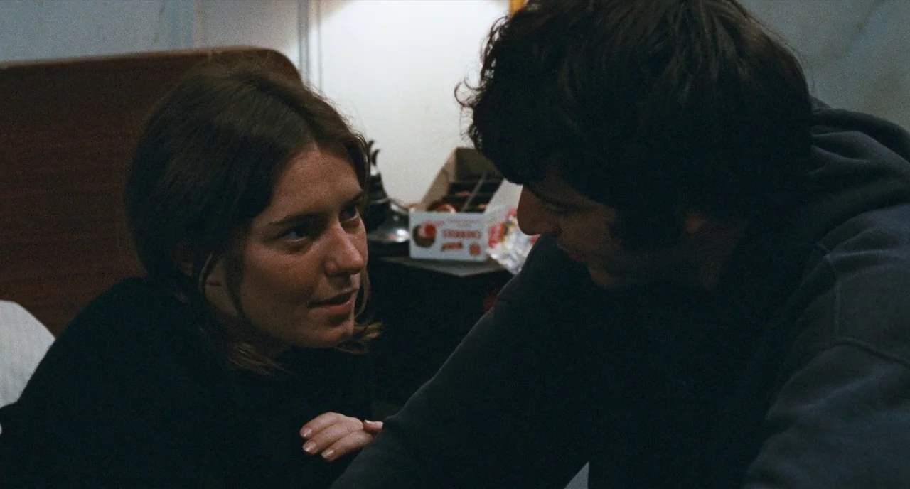 The Panic in Needle Park (1971) movie still: medium — A dark room with a wooden headboard and a box on a side table. A man and woman sit facing each…