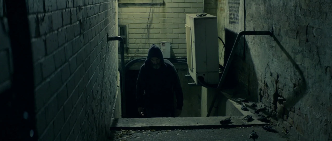 You Were Never Really Here (2017) movie still: medium — A dark, narrow stairwell is flanked by two rough-textured brick walls. A bearded man in a…