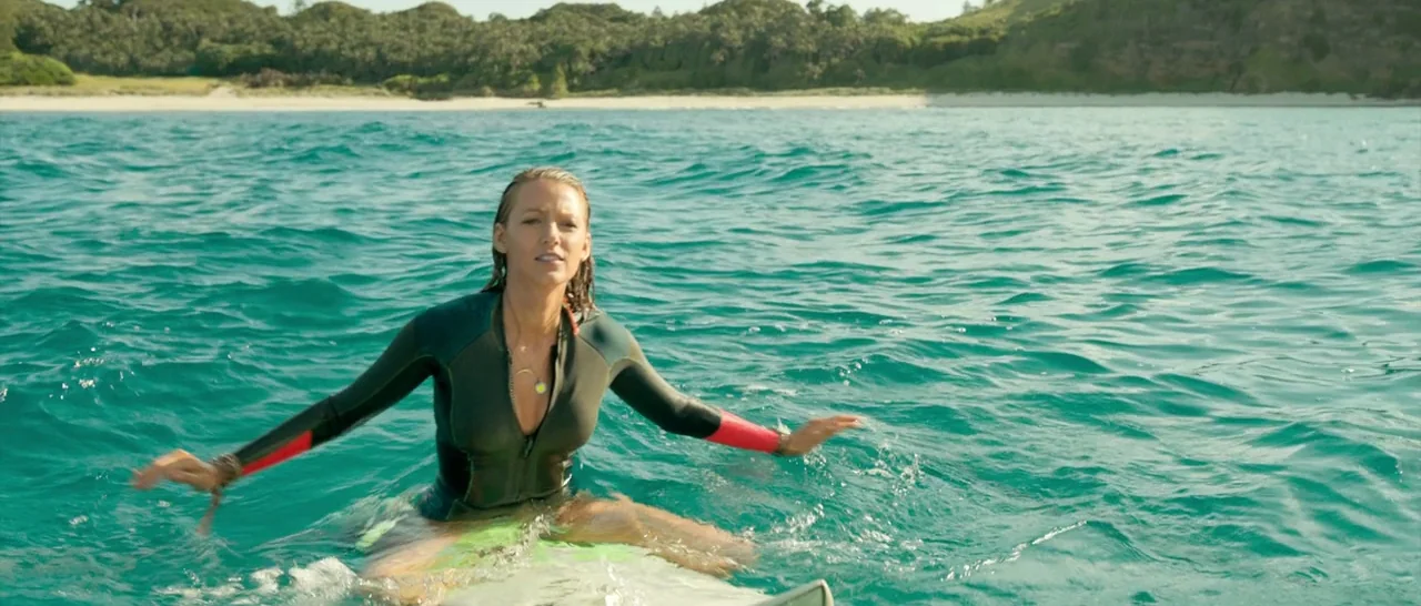 The Shallows (2016) movie still: medium — A turquoise ocean surface with a distant sandy beach and dense green coastline vegetation. A young woman…