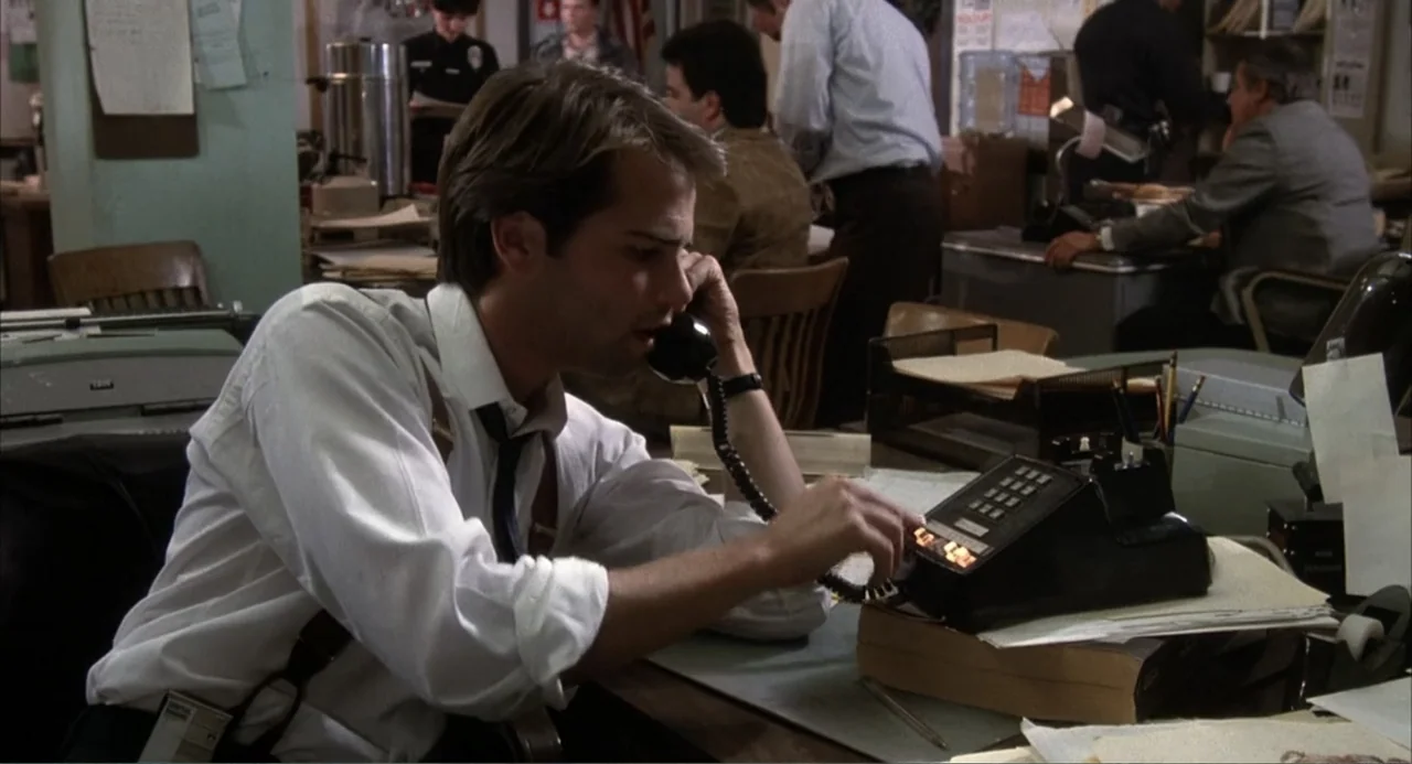 I, Madman (1989) movie still: medium — A crowded 1980s police department office features cluttered desks and several uniformed personnel. A man sits…