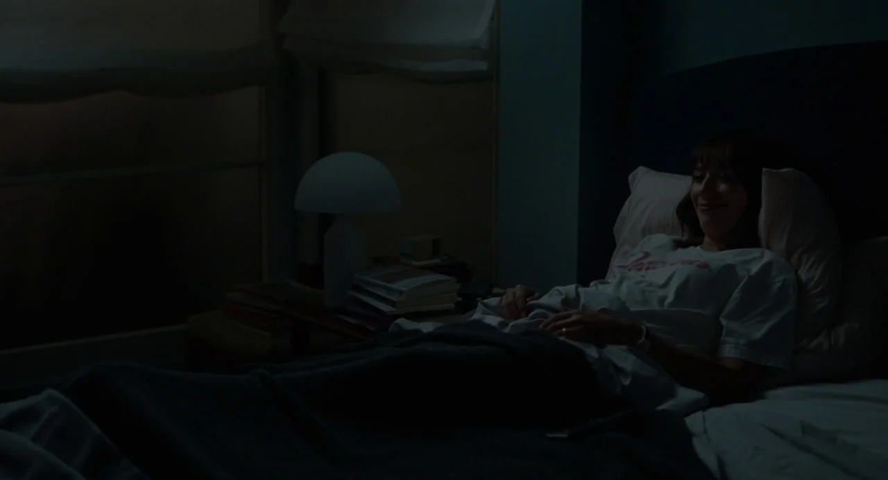On The Rocks (2020) movie still: medium — A dimly lit room contains a bed with a person and a bedside table. A woman lies in bed, gazing toward the…
