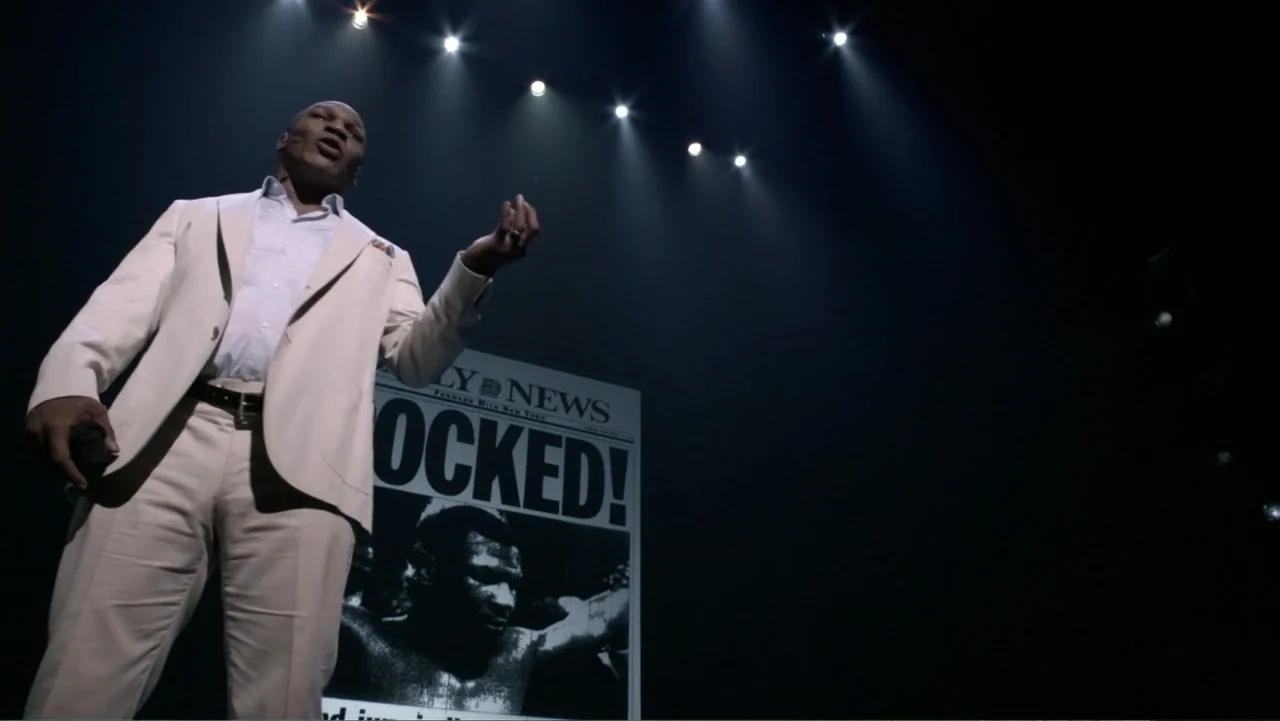 Mike Tyson: Undisputed Truth (2013) movie still: fullbody — A dark stage features a single man standing before a large newspaper prop display. The…
