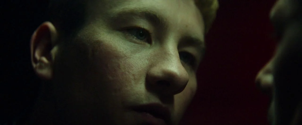 Calm With Horses (2019) movie still: closeup — A dark, dimly lit environment features a person obscured in shadows to the right. A young man with…