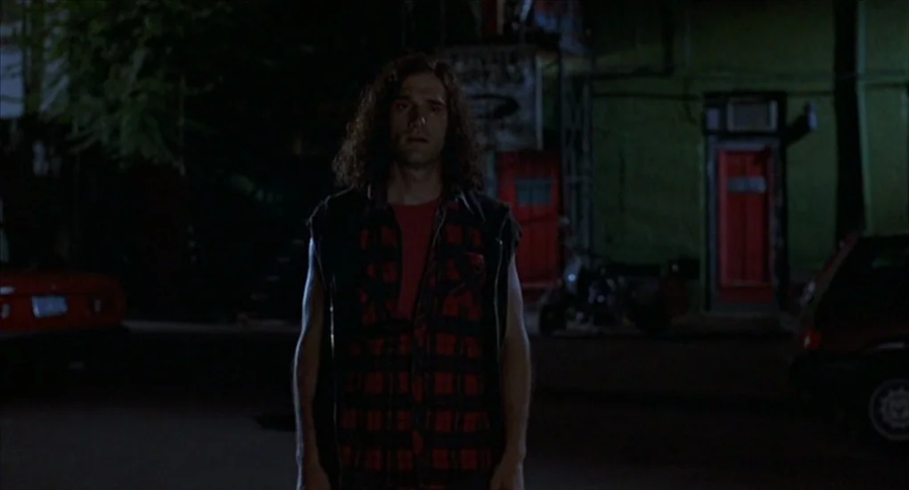Exotica (1994) movie still: medium — A night parking lot with a low-rise building in the background featuring red doors. A young man with long hair…