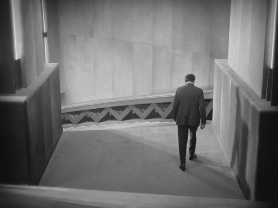 Metropolis (1927) movie still: fullbody — A modernist staircase features concrete walls with a repetitive zig-zag railing pattern. A man in a dark…