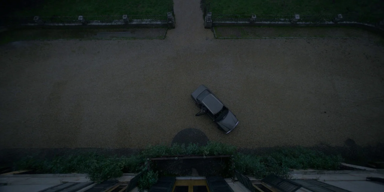 Immaculate (2024) movie still: establishing — A gravel courtyard is situated in front of a building with an open doorway. A dark grey sedan is…
