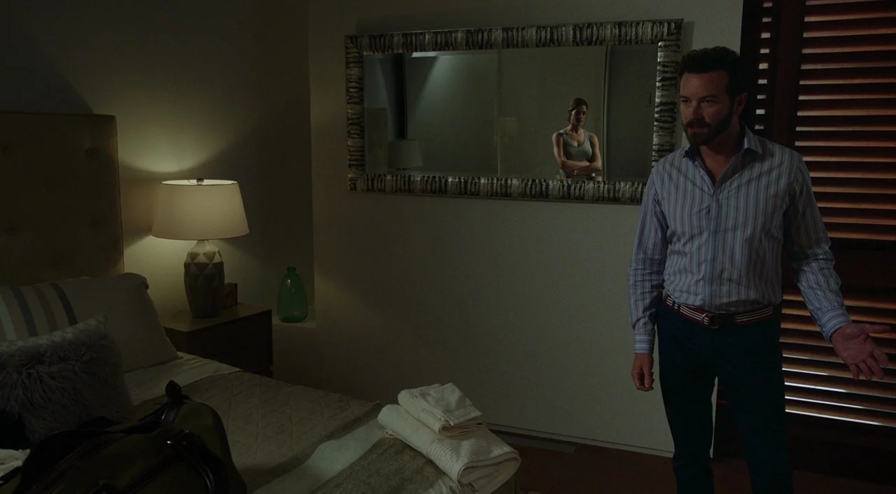 Urge (2016) movie still: medium — A hotel room features a lamp on a nightstand and a wide mirror on wall. A man stands in the foreground while a…