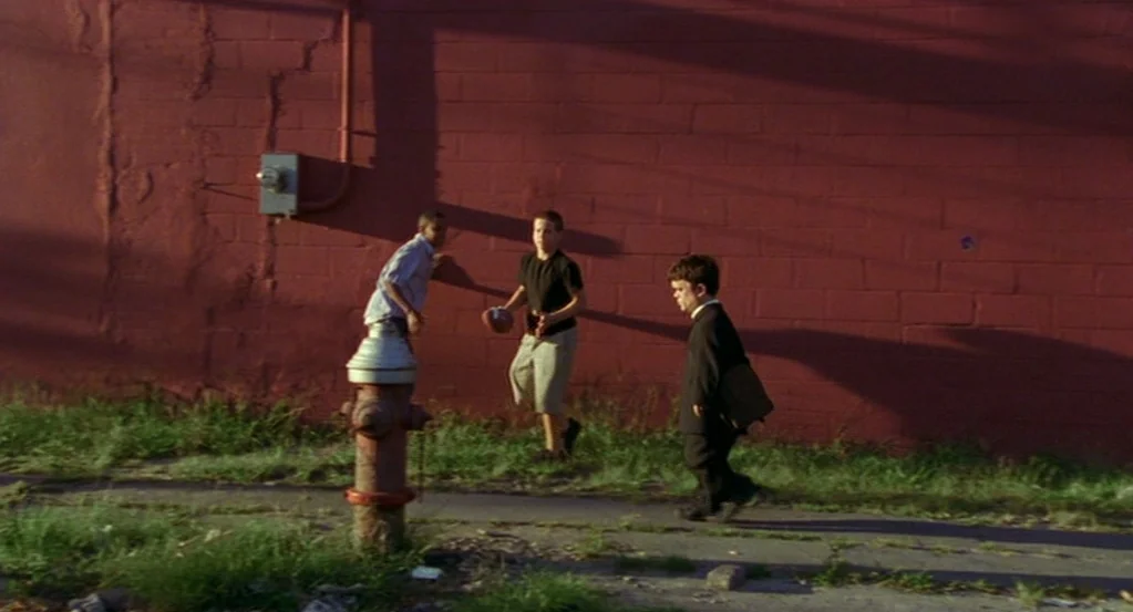 The Station Agent (2003) movie still: wide — A sidewalk runs parallel to a long, painted red brick wall in the evening light. Three people occupy…